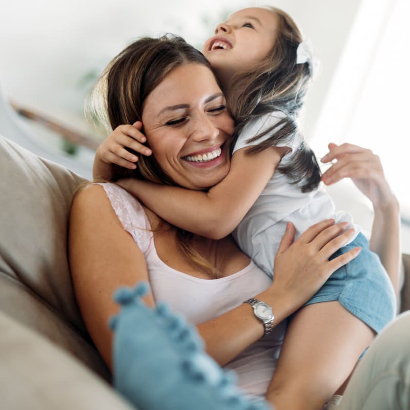 A smiling woman sitting on a beige couch while her young daughter embraces her in a big, happy hug.