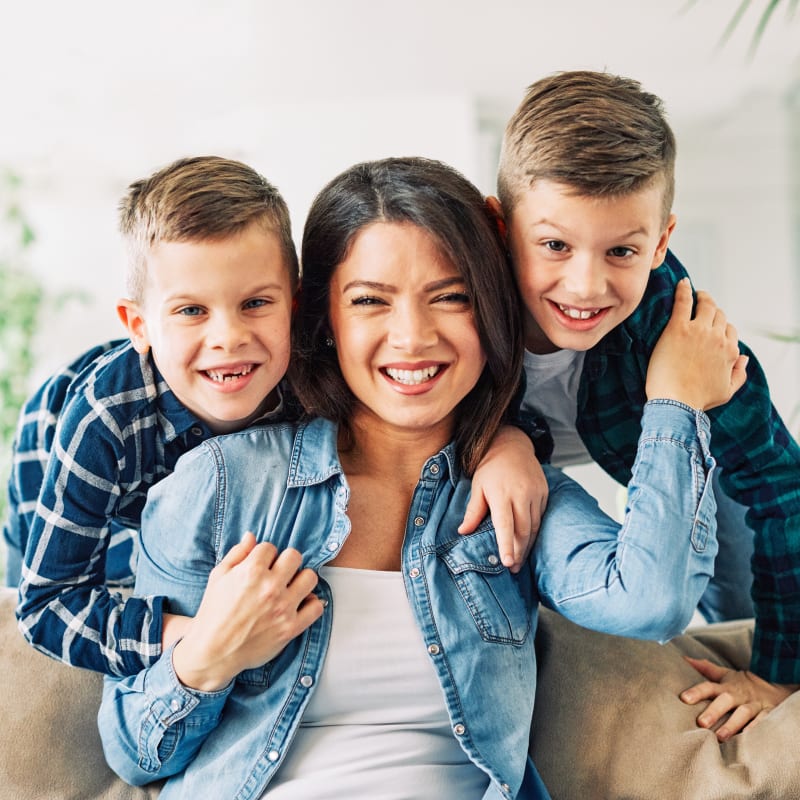 A woman sits while two young boys hug her from behind. They are all smiling.