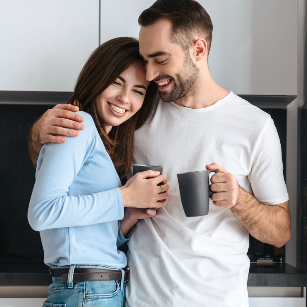 A man stands with an arm around a woman in a kitchen while they each hold a coffee mug.