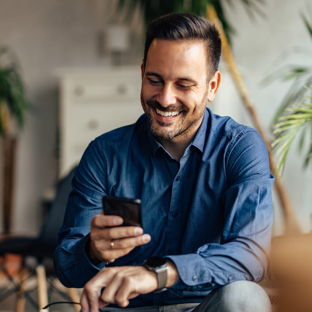 A man is sitting down in a room with plants behind him. He is looking at his phone and smiling.