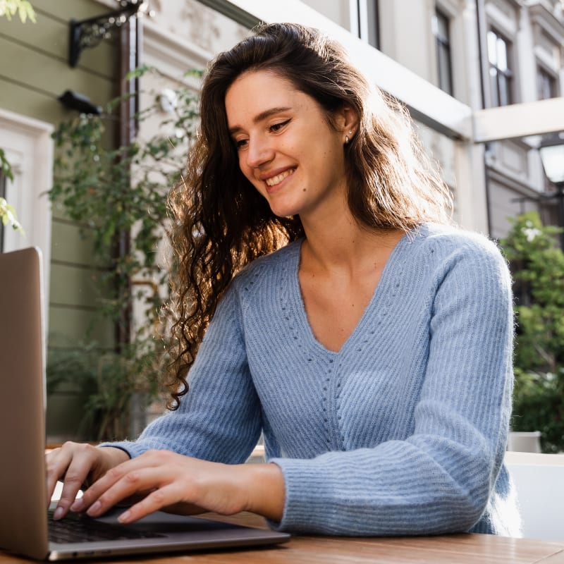 A woman is sitting outside at a table with trees behind her while she looks at a laptop.