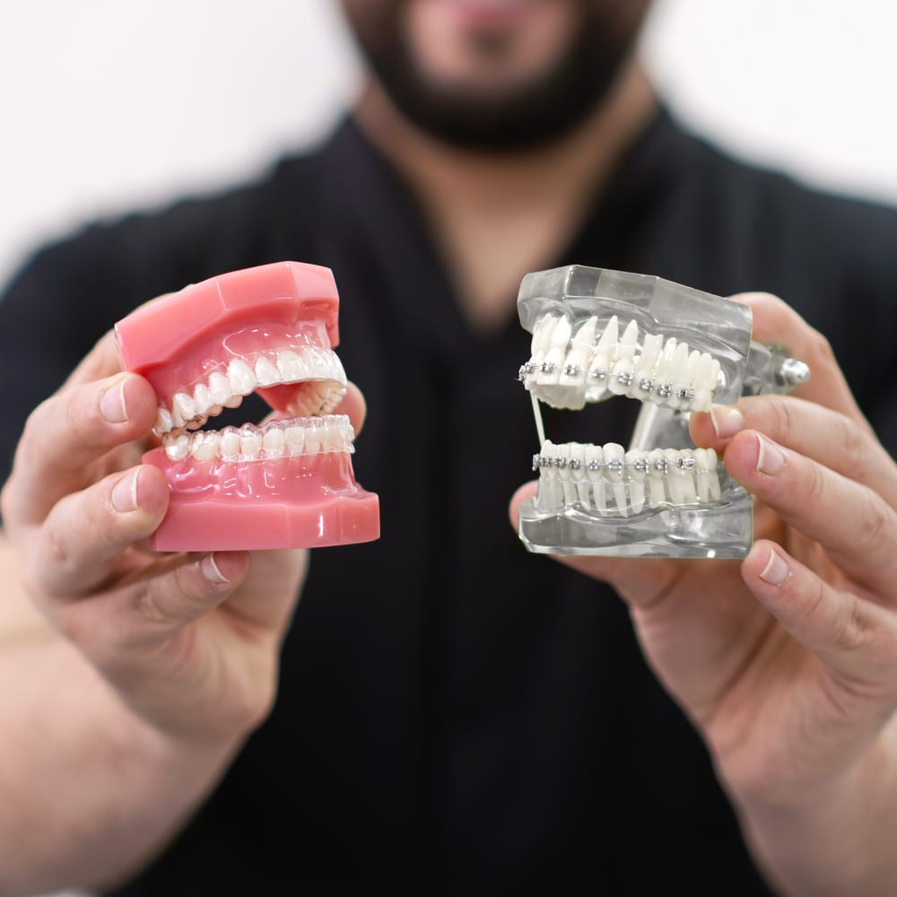 A dentist hols up an example of a mouth with Invisalign clear aligners in, and a set of teeth with traditional braces.