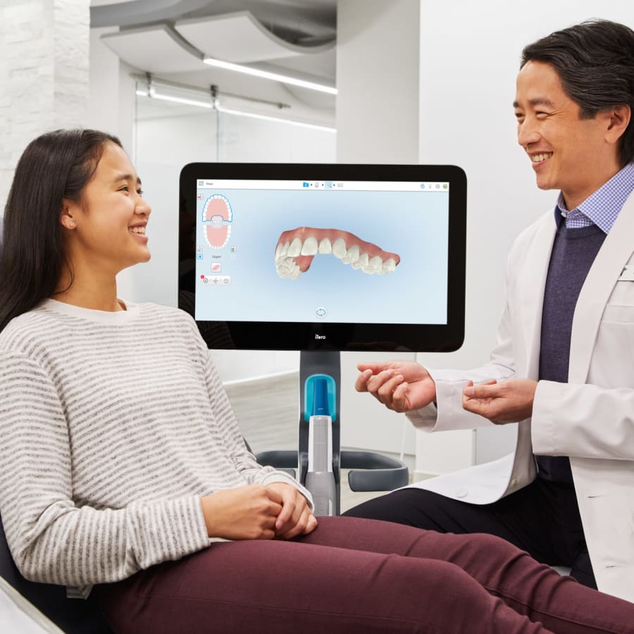 A dentist and a patient review the patient's oral health. A screen shows a digital rendering of her upper teeth.