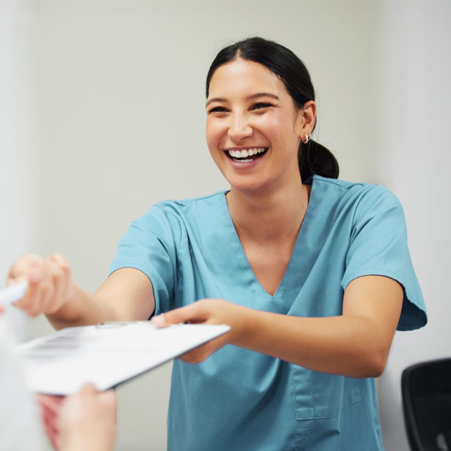 A dental assistant is smiling brightly as she passes someone a clipboard.