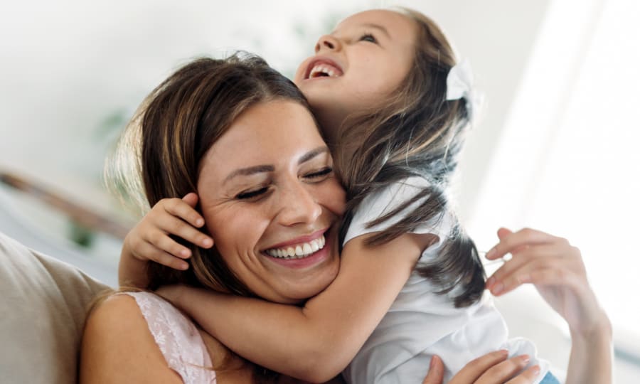 Woman with straight white teeth is smiling while being hugged by a little girl.