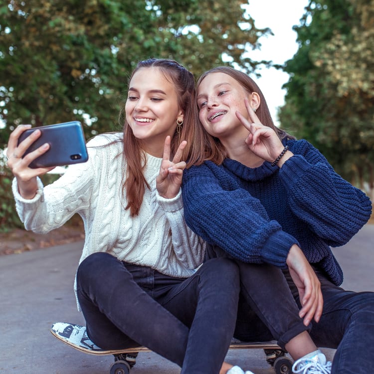 Two teen girls are sitting on a skateboard outside taking a selfie while holding up peace signs.